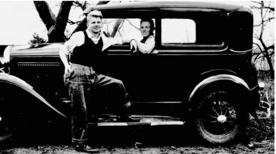 Vintage photo of old car with one person sitting inside and one person standing on the running boards